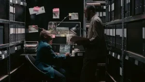 Two investigators reviewing case files and surveillance photos in a secure office filled with organised binders and evidence boards.